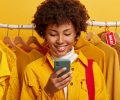 Glad online merchant focused in smartphone device, enjoys biggest shopping day, rejoices marked down prices, stands against yellow clothing on racks, celebrates importnat ecommerce holiday in autumn