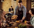 Group of happy friends having fun while waiter is serving them food in a pub. Focus is on waiter.