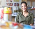 Picture of charming charismatic young female artist wearing khaki shirt grinning broadly feeling happy about her job and creating process, sitting at workshop, surrounded with painting accessories