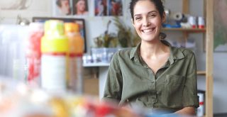 Picture of charming charismatic young female artist wearing khaki shirt grinning broadly feeling happy about her job and creating process, sitting at workshop, surrounded with painting accessories