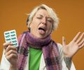 sick unhealthy woman with short hair in warm scarf and hat holding blister with pills raising arm sticking out tongue with disgusted expression standing over orange background